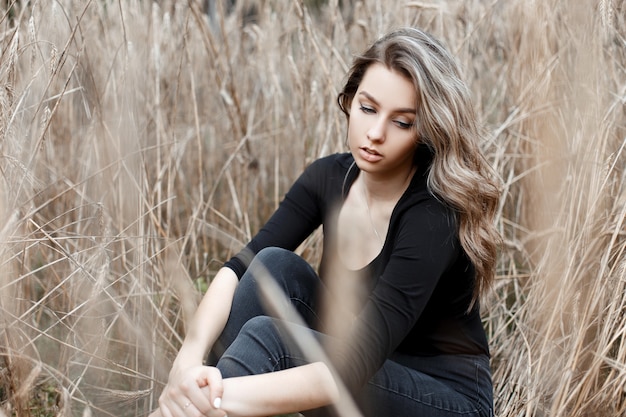 Uma jovem mulher muito fofa em jeans elegantes e uma camiseta preta da moda está descansando sentada no chão no campo