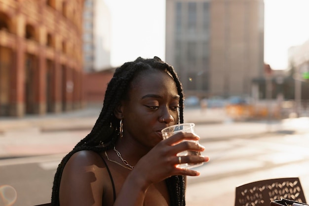 Uma jovem negra com tranças no cabelo bebendo um refrigerante sentada em um terraço de uma cafeteria na cidade