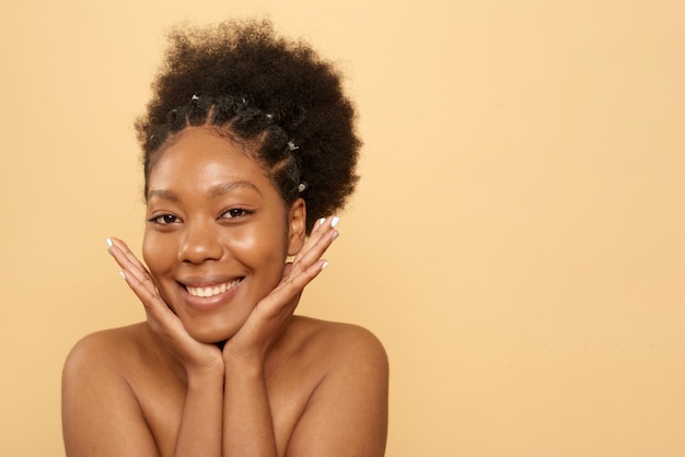 Uma jovem negra positiva com ombros nus e pele bonita tocando o rosto olhando para a câmera no fundo bege