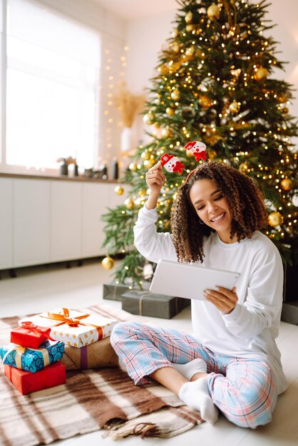 Uma jovem no fundo da árvore de Natal com presentes com um tablet tem chamada de vídeo ou bate-papo por vídeo.