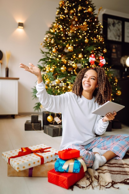 Uma jovem no fundo de uma árvore de Natal com presentes com um tablet faz uma videochamada.