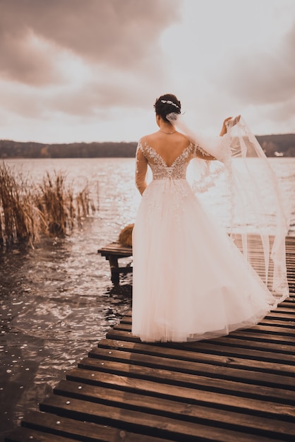 Uma jovem noiva com cabelo ruivo cacheado vestido de noiva branco fica no cais de madeira marrom no meio do lago azul-celeste.