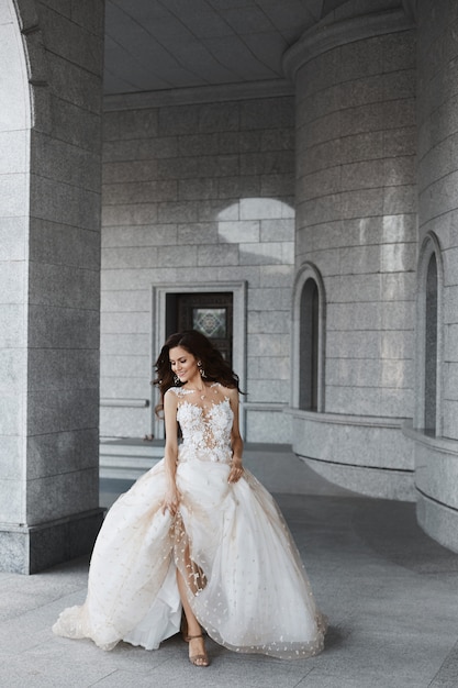 Uma jovem noiva feliz com um penteado de casamento em um vestido de renda branca atravessa o pátio de uma igreja antiga.