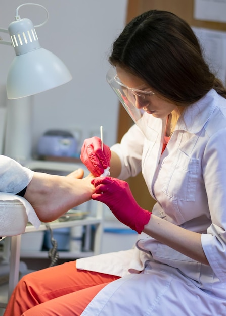 Uma jovem podóloga com tela protetora faz uma pedicure em um paciente durante uma pandemia