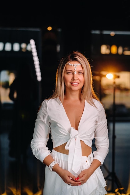 Uma jovem positiva em um vestido branco e um ornamento na cabeça sorri para a câmera enquanto está no estúdio