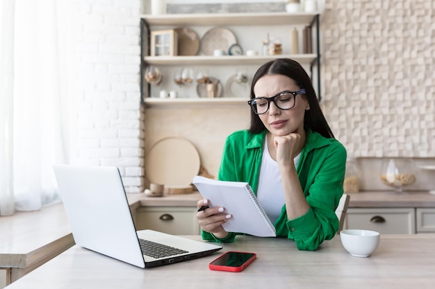 Uma jovem preocupada senta-se em casa na cozinha com um laptop e anota o orçamento familiar em um