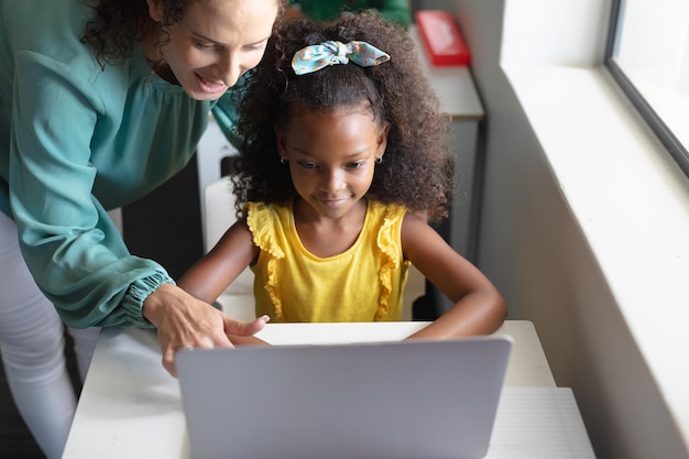 Uma jovem professora caucasiana sorridente a ensinar computador a uma rapariga afro-americana na secretária.