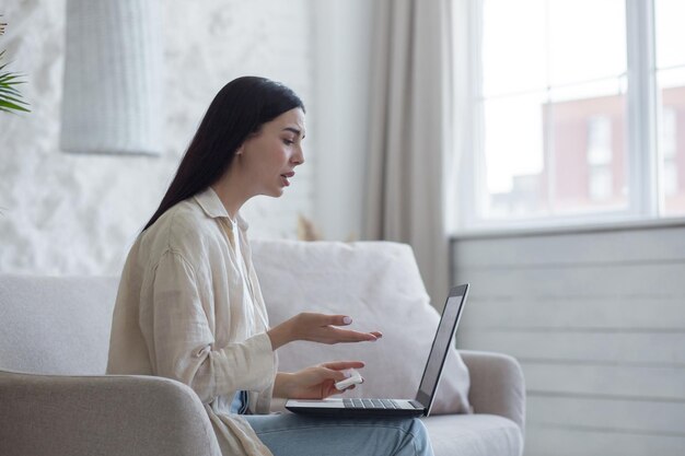 Uma jovem refugiada chateada senta em um sofá em um apartamento e fala em uma videochamada de um laptop