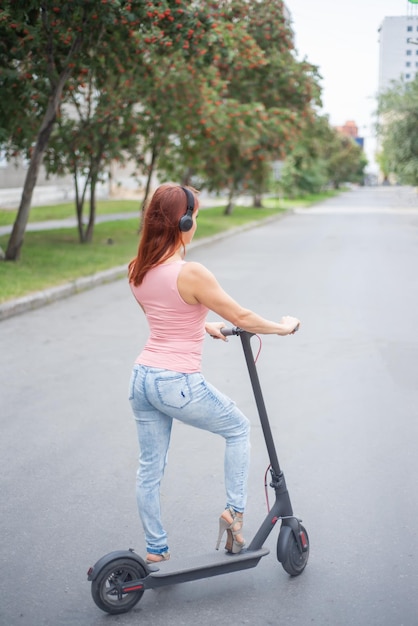 Uma jovem ruiva em jeans rasgados e sandálias de salto alto anda de scooter elétrica na estrada e ouve música em seus fones de ouvido sem fio bluetooth Transporte elétrico conveniente