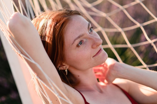 Uma jovem ruiva sem maquiagem está descansando em uma rede em um campo de lavanda Férias de verão e tempo de viagem