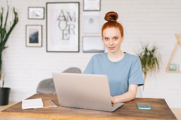 Uma jovem ruiva sorridente e feliz trabalhando em um laptop na mesa do escritório em casa