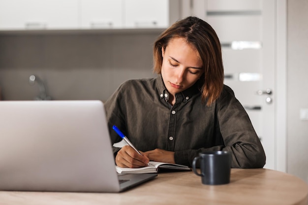 Uma jovem se senta a uma mesa e escreve em um caderno, olhando para um laptop