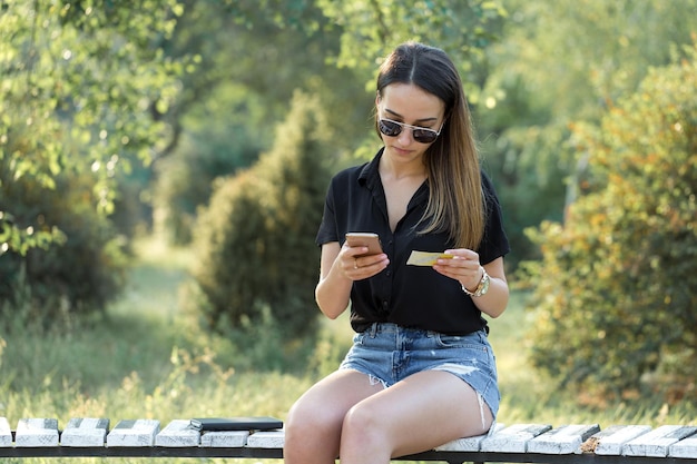 Uma jovem se senta em um banco em um parque e faz anotações Vestida em um estilo livre Mulher de negócios falando ao telefone com clientes