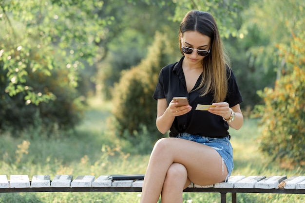 Uma jovem se senta em um banco em um parque e faz anotações Vestida em um estilo livre Mulher de negócios falando ao telefone com clientes