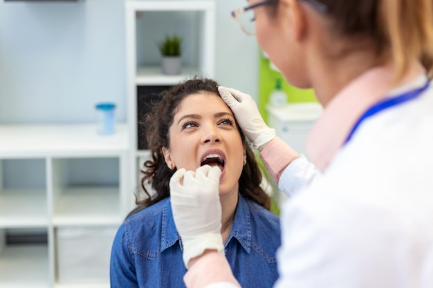 Uma jovem se senta em uma mesa de exame em frente ao médico O médico estende a mão com um abaixador de língua enquanto a mulher olha para cima e mostra a língua