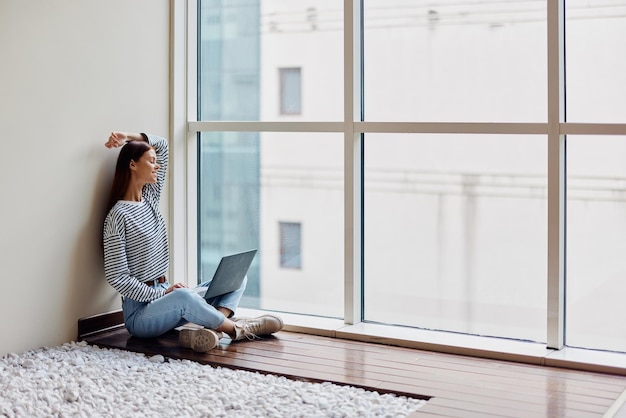 Uma jovem se senta no chão com seu laptop perto da janela trabalhando e estudando na cidade grande