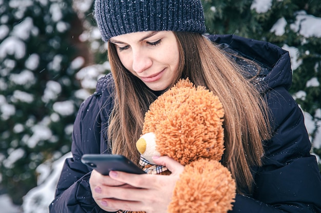 Uma jovem segura um ursinho de pelúcia e um smartphone nas mãos em tempo de neve