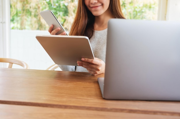 Uma jovem segurando e usando um telefone celular, um tablet pc e um laptop em uma mesa de madeira