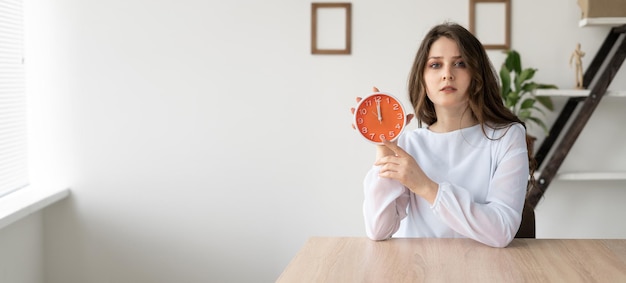 Uma jovem senhora caucasiana senta-se em casa em uma mesa e segura um relógio nas mãos São 12 horas da tarde para o almoço Descansar e descansar durante o banner do dia de trabalho