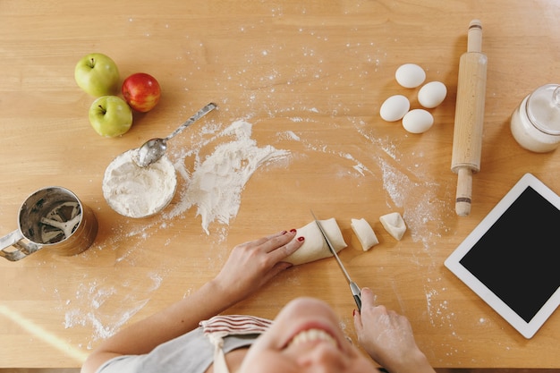 Uma jovem sentada à mesa com um tablet corta uma massa com uma faca em pedaços em casa na cozinha. Cozinhando em casa. Preparar comida. Vista do topo.