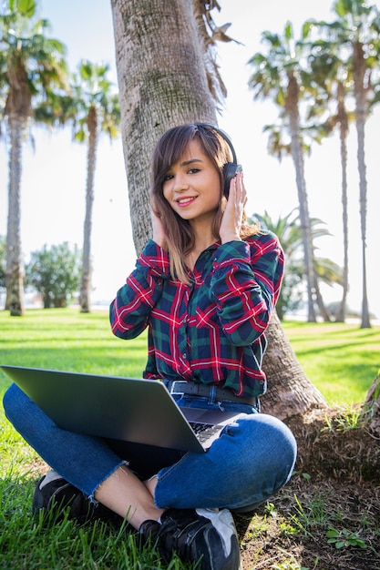 Uma jovem sentada em um parque público usa seu laptop e ouve áudio em seus fones de ouvido