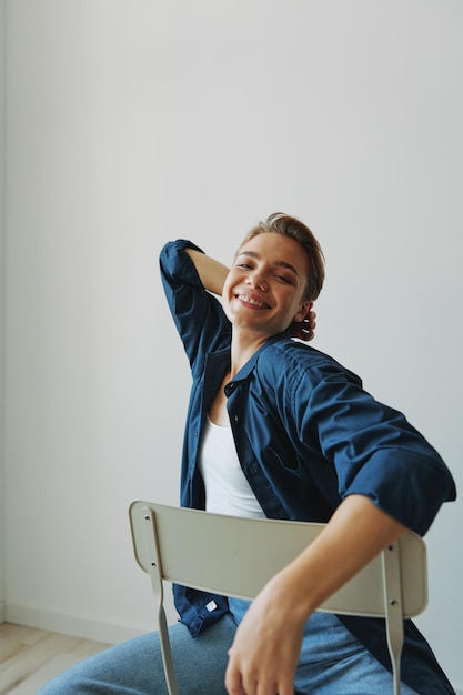 Uma jovem sentada em uma cadeira em casa sorrindo com os dentes com um corte de cabelo curto em jeans e uma camisa jeans em um fundo branco Menina poses naturais sem filtros