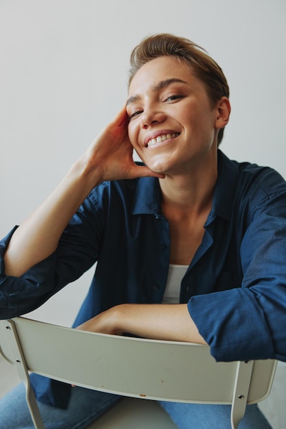 Uma jovem sentada em uma cadeira em casa sorrindo com os dentes com um corte de cabelo curto em jeans e uma camisa jeans em um fundo branco Menina poses naturais sem filtros