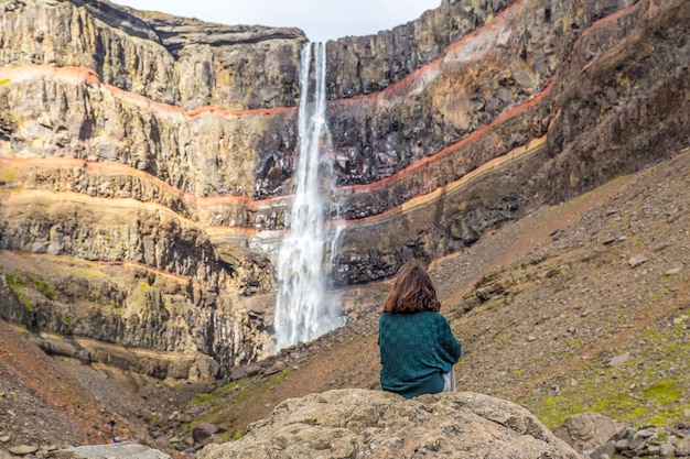 Uma jovem sentada em uma das maravilhas da Islândia, a cachoeira Hengifoss