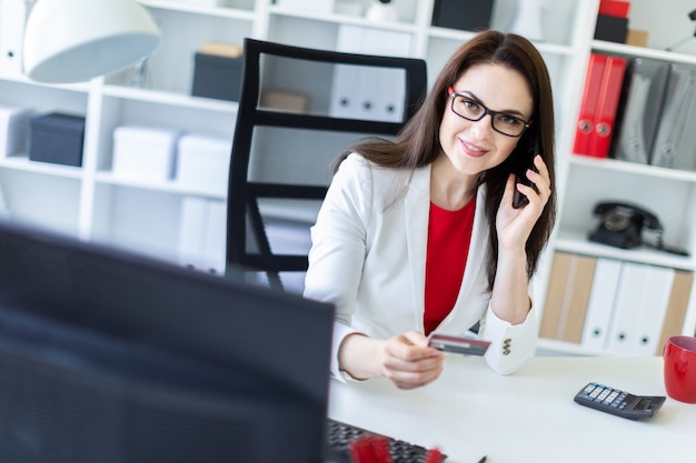 Uma jovem sentada no escritório à mesa e segurando um cartão e telefone bancário.