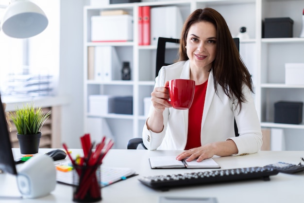 Uma jovem sentada no escritório, na mesa do computador e segurando uma xícara vermelha.