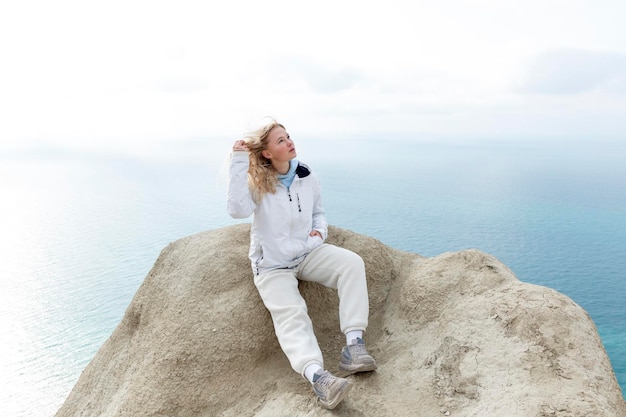 Foto uma jovem sentada no topo de uma montanha acima do mar em um dia ensolarado loira bonita com um casaco branco e calças de treino estilo de vida ativo turismo e viagens