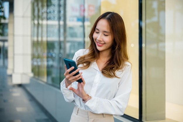 Uma jovem sorri enquanto usa seu smartphone ao ar livre Ela segura o dispositivo com uma mão e digita com a outra A mulher está feliz e conectada