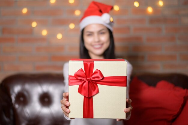 Foto uma jovem sorridente com chapéu de papai noel vermelho e mostrando uma caixa de presente no dia de natal