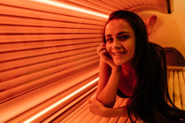 Uma jovem sorridente deitada em uma cama de bronzeamento artificial sob os raios ultravioleta, tomando banho de sol, quer uma pele bronzeada