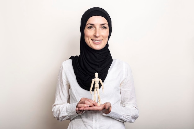 Uma jovem sorridente em um hijab segura um fantoche de um homem de madeira sobre um fundo claro