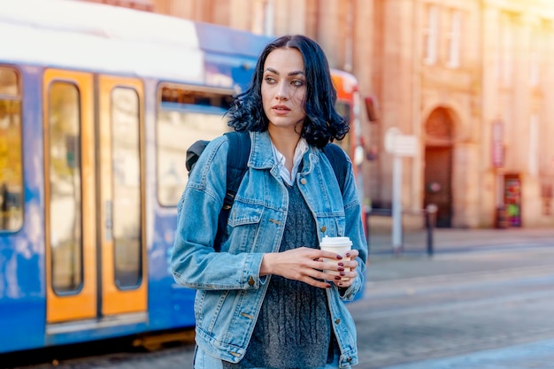 Uma jovem sorridente em uma jaqueta jeans tomando café e esperando um bonde na parada