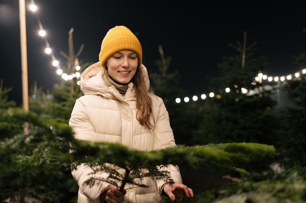 Uma jovem sorridente escolhe galhos de árvores de Natal para decorar a casa para os feriados de Ano Novo contra o fundo de luzes brilhantes