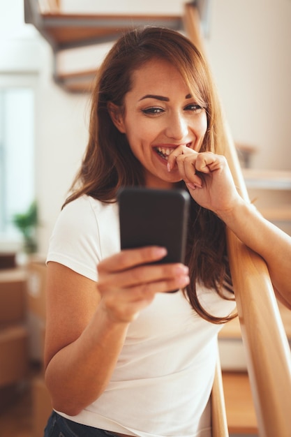 Uma jovem sorridente feliz fica na sala de seu novo apartamento em frente às escadas olhando para smartphone.