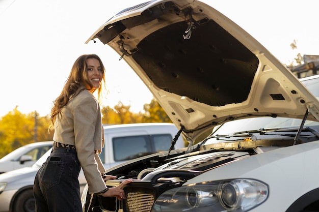 Uma jovem sorridente fica perto de um capô aberto de um carro O conceito de avaria do serviço de carro carro quebrado