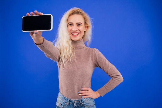 Uma jovem sorridente segura uma tela de telefone para a câmera em um fundo azul Mockup