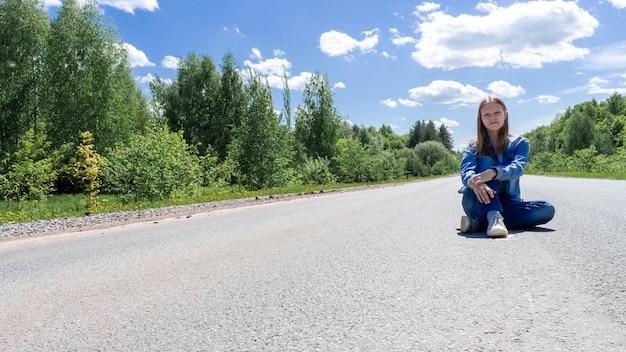 Uma jovem sorridente, uma adolescente, senta-se no meio da estrada no verão fora da cidade