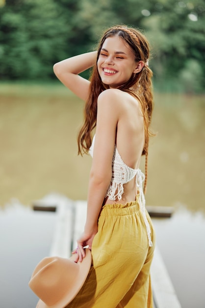 Uma jovem sorrindo em uma imagem de um hippie e ecodress dançando na natureza à beira do lago usando um chapéu e calças amarelas no pôr do sol de verão