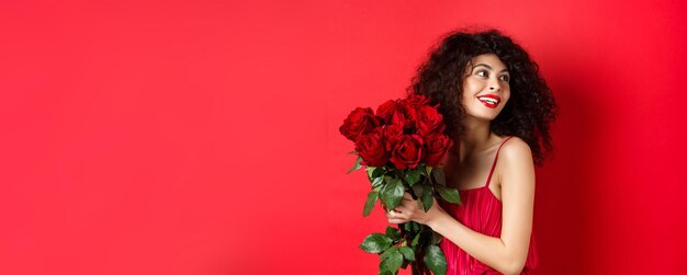 Uma jovem tenra com um elegante vestido vermelho segurando um buquê romântico de rosas vermelhas e parecendo sorridente