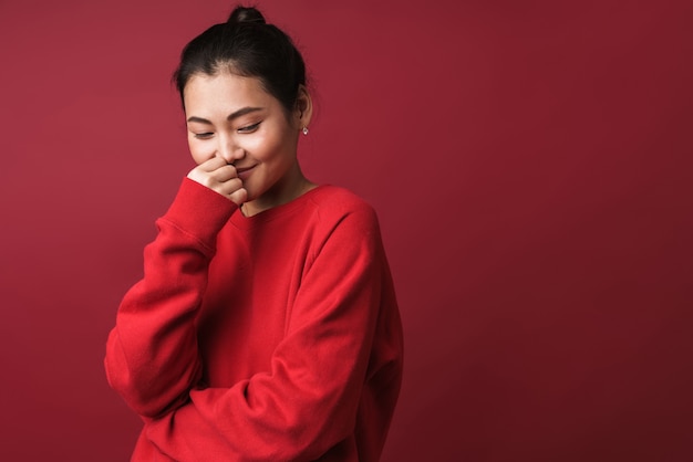 uma jovem tímida sorridente asiática posando isolada sobre uma parede vermelha.