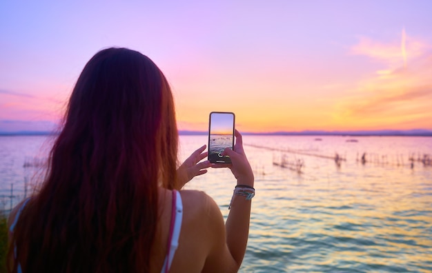 Uma jovem tirando uma foto com o celular em uma paisagem ao pôr do sol. Albufera de Valência. Espanha