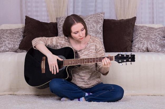 Uma jovem toca um violão preto no tapete da sala