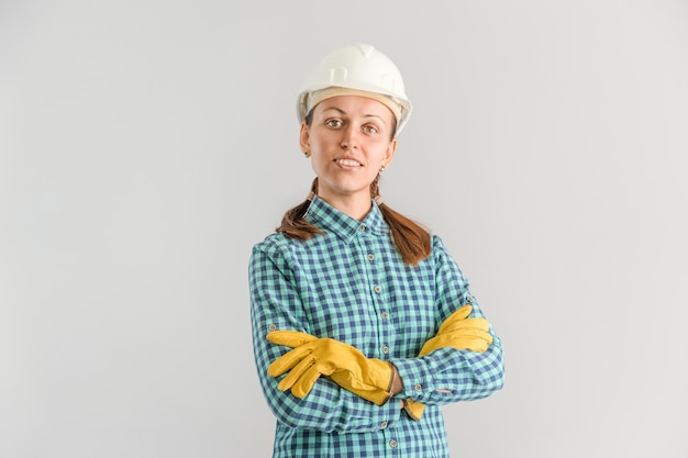 Uma jovem trabalhadora da construção civil adulta com um capacete branco de construção em pé com os braços cruzados sobre um fundo cinza