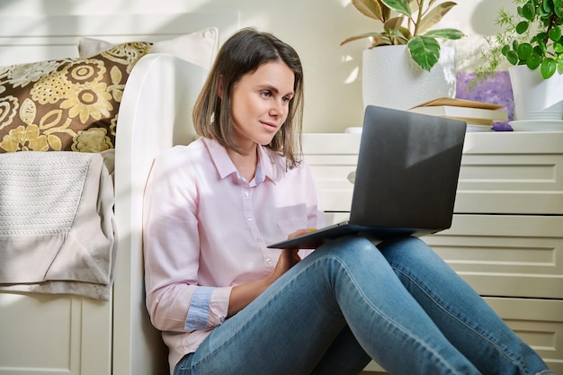 Uma jovem triste e séria olhando para a tela do laptop sentada no chão em casa