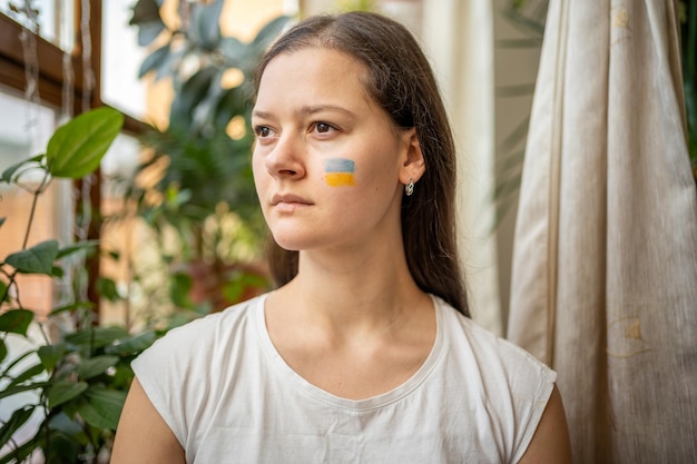 Uma jovem ucraniana triste com a bandeira da ucrânia no rosto está olhando pela janela o conceito