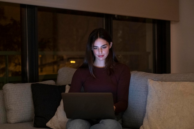 Uma jovem usando laptop à noite em casa sentada no sofá pesquisando na web e trabalhando até tarde As mulheres usam tecnologia de computador de conexão sem fio para trabalho ou atividade interior de lazer